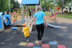 Blonde woman with blue shirt holding hands of child in yellow coat walking away from the camera towards a playground