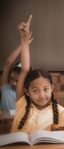 Girl in yellow shirt with pigtail braids raising her hand to ask a question in class