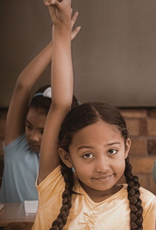 Girl in yellow shirt with pigtail braids raising her hand to ask a question in class