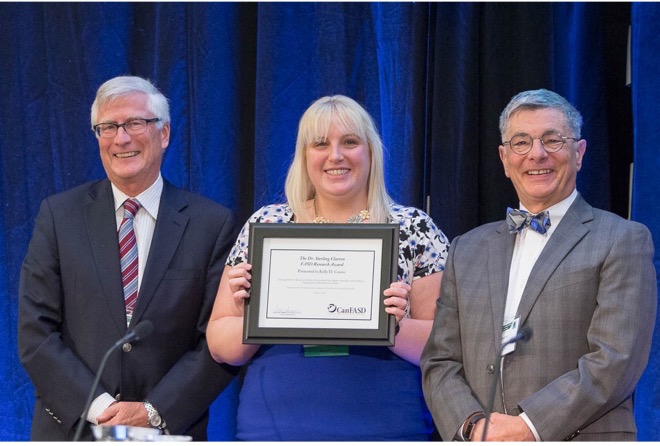 Kelly holding a framed certificate of her award and smiling at the camera with Dr. Sterling Clarren standing and smiling to her left and Dr. Alan Bocking standing and smiling on her right.