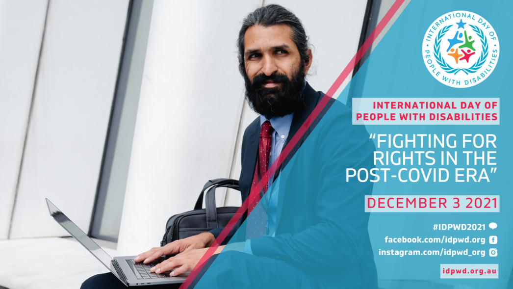 Man in a suit with a beard is sitting, working on his laptop. He turns to smile to someone behind the camera.