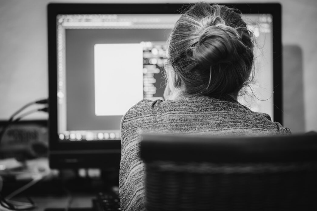 Black and white image taken from behind of a woman with a bun sitting in a chair and working on the computer.
