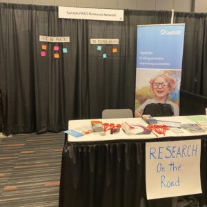 Black conference booth with 'Research on the Road' sign. There is a large banner in the background and colourful sticky notes stuck to the back of the booth.