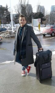 RJ smiling at the camera holding a suitcase. He is standing on the sidewalk with his feet crossed, wearing red sneakers, jeans, and a grey coat.