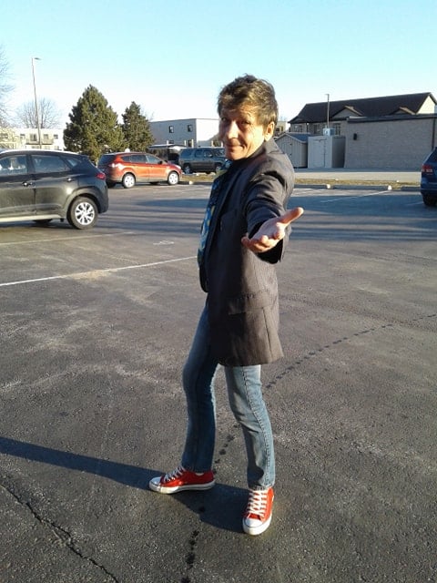 RJ Formanek in a parking lot gesturing at the camera with an open hand for you to follow him. He is dressed in a grey coat, red sneakers, and jeans. His body is angled away from the camera but his head is turned back to face the lens. 