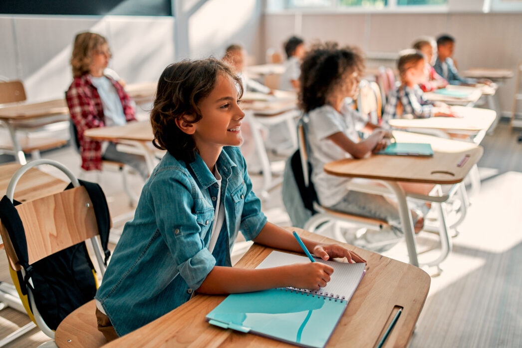 Children sitting in desks smiling and looking out of the frame at the front of the classroom. They are ready to study with their pen on the paper in front of them.