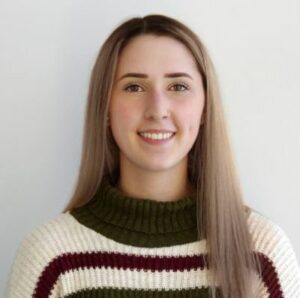 Kathleen smiles at the camera wearing a striped sweater and standing in front of a white wall