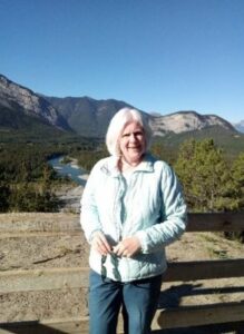 Lyn Penfold poses for the camera, smiling, in front of mountains in the summer. 
