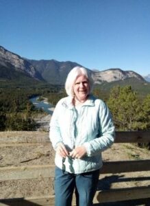 Lyn Penfold poses for the camera, smiling, in front of mountains in the summer.