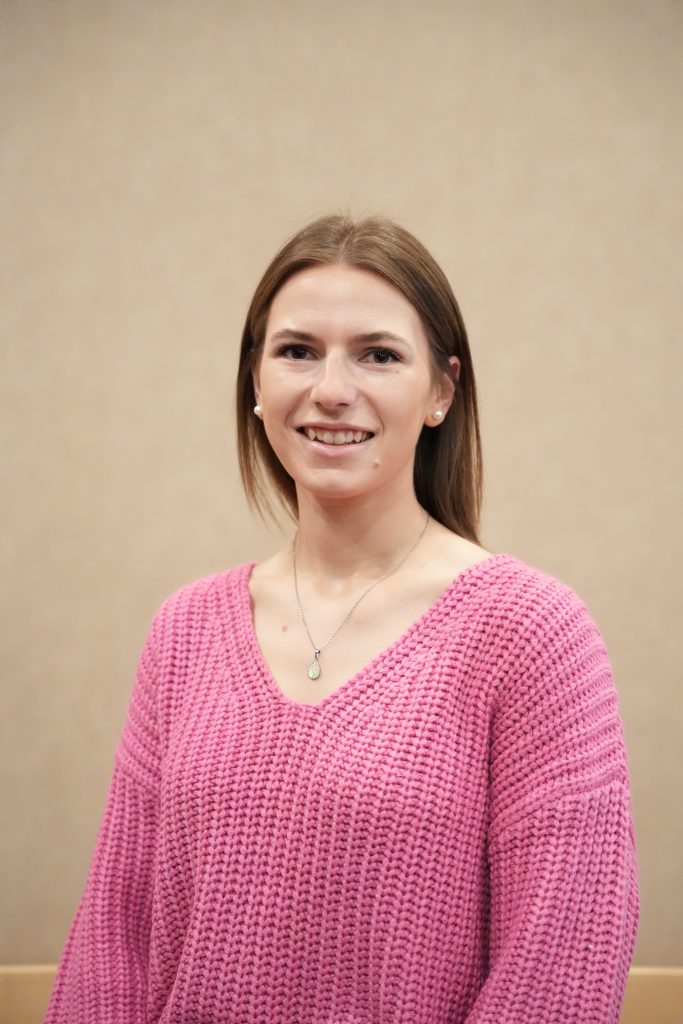 Kirsten Morrison headshot A woman with straight, light brown hair smiles at the camera. She is wearing a bright pink knit sweater, pearl earrings, and a pendant necklace, standing in front of a plain beige background.
