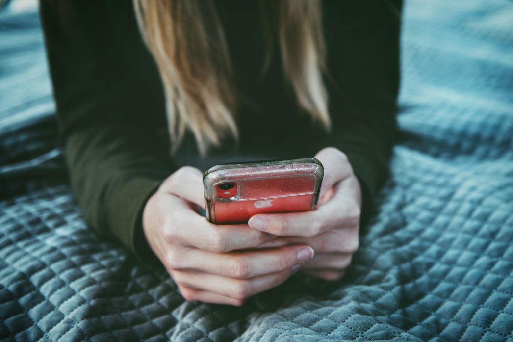 Close-up of a person with long blond hair, wearing a dark sweater, sitting on a quilted blanket and holding a red iPhone with a clear case, focused on the phone screen.