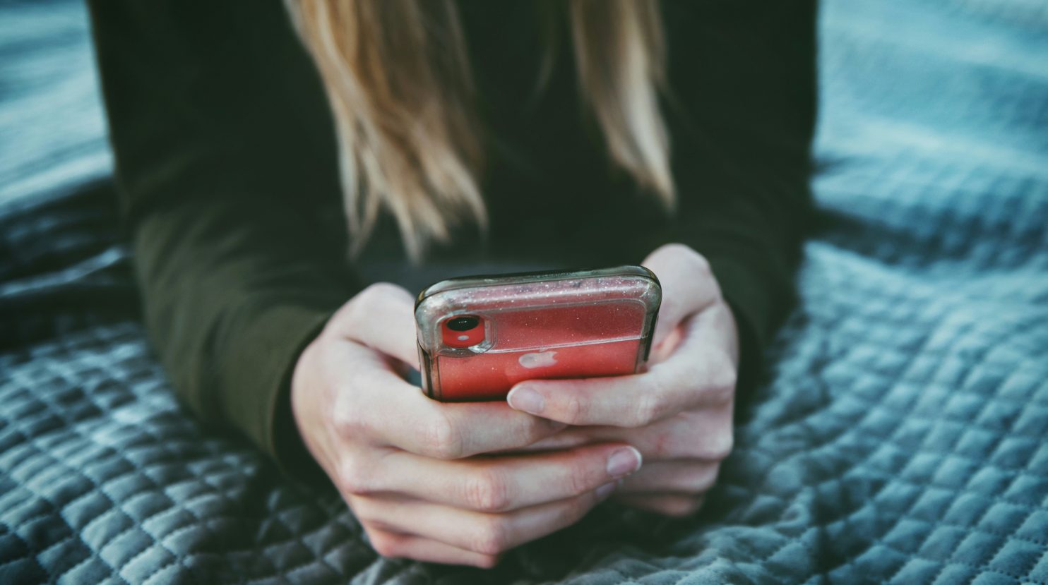 Close-up of a person with long blond hair, wearing a dark sweater, sitting on a quilted blanket and holding a red iPhone with a clear case, focused on the phone screen.