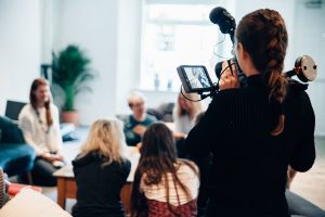 A filmmaker with a braided hairstyle is capturing a group discussion using a professional video camera with a monitor and microphone. The setting is a bright, modern indoor space with natural light, a wooden table, and a couch. Several people are engaged in conversation in the background, slightly out of focus.