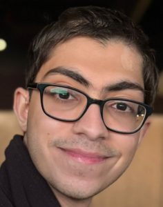 A young man with short dark hair, wearing black rectangular glasses and a black jacket, smiling at the camera in a warmly lit indoor setting.