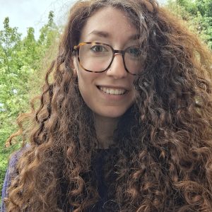 A young woman with long, curly brown hair and tortoiseshell glasses smiles at the camera. She is outdoors, with green trees in the background, and is wearing a dark top.