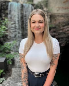 A woman with long straight blonde hair stands in front of a stone waterfall.