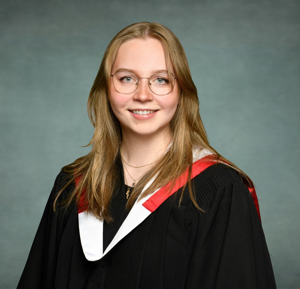 A young woman wearing graduation robes with a black gown and a red and white stole. She has long, light brown hair, is wearing glasses, and is smiling at the camera. The background is a solid, muted blue-grey color.