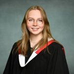 A young woman wearing graduation robes with a black gown and a red and white stole. She has long, light brown hair, is wearing glasses, and is smiling at the camera. The background is a solid, muted blue-grey color.
