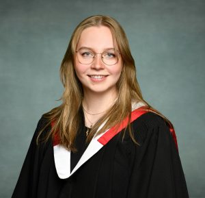 A young woman wearing graduation robes with a black gown and a red and white stole. She has long, light brown hair, is wearing glasses, and is smiling at the camera. The background is a solid, muted blue-grey color.