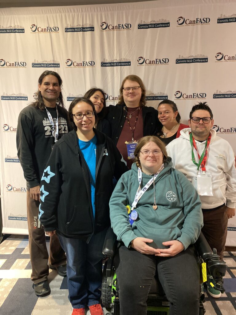 A group of eight adults pose together in front of a step-and-repeat banner for the Canada FASD Conference. One person in front is seated in a power wheelchair, while the others stand closely behind and beside them. The group is smiling, wearing conference lanyards and casual clothing.